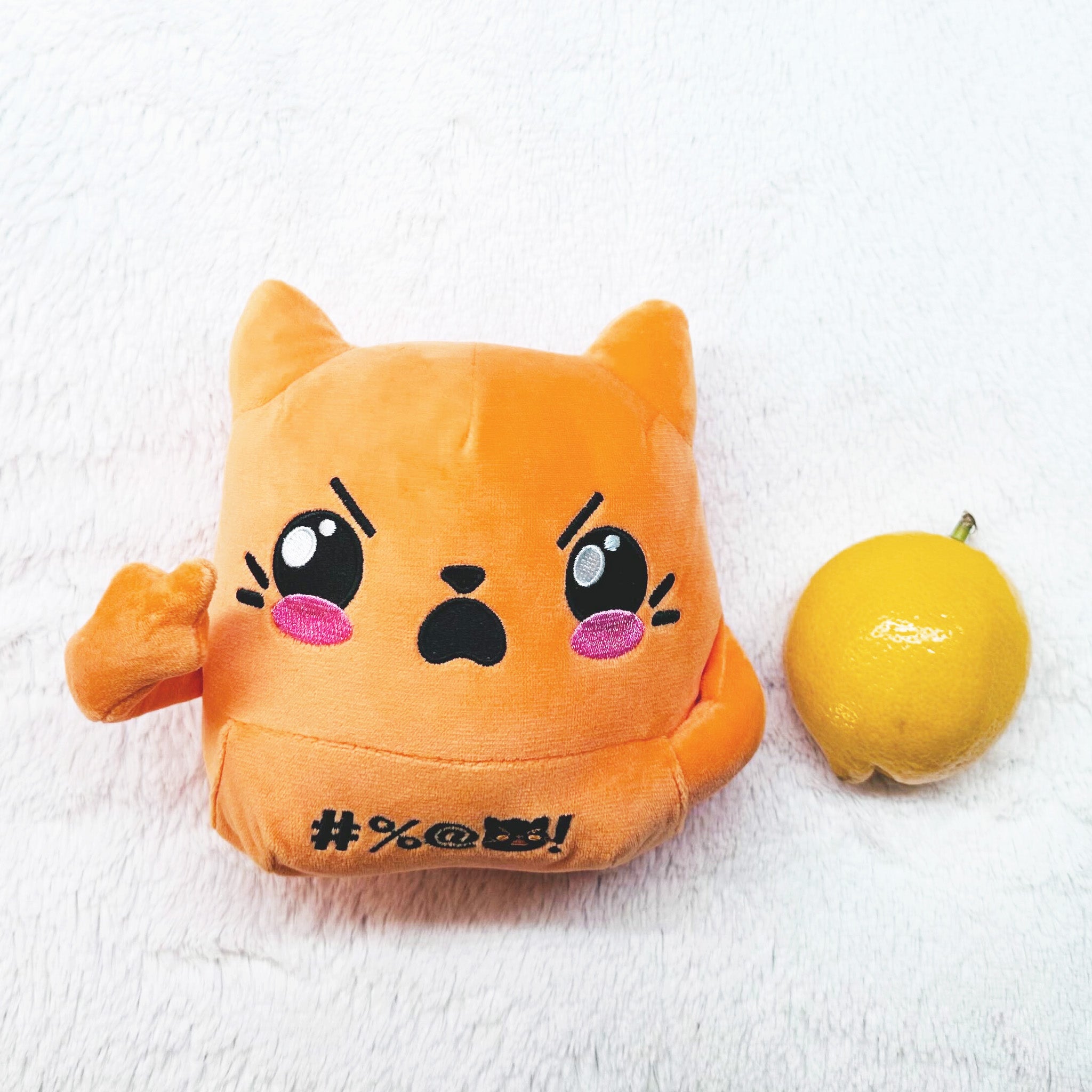 Orange cat plush, one middle finger raised, lying face up next to a lemon.