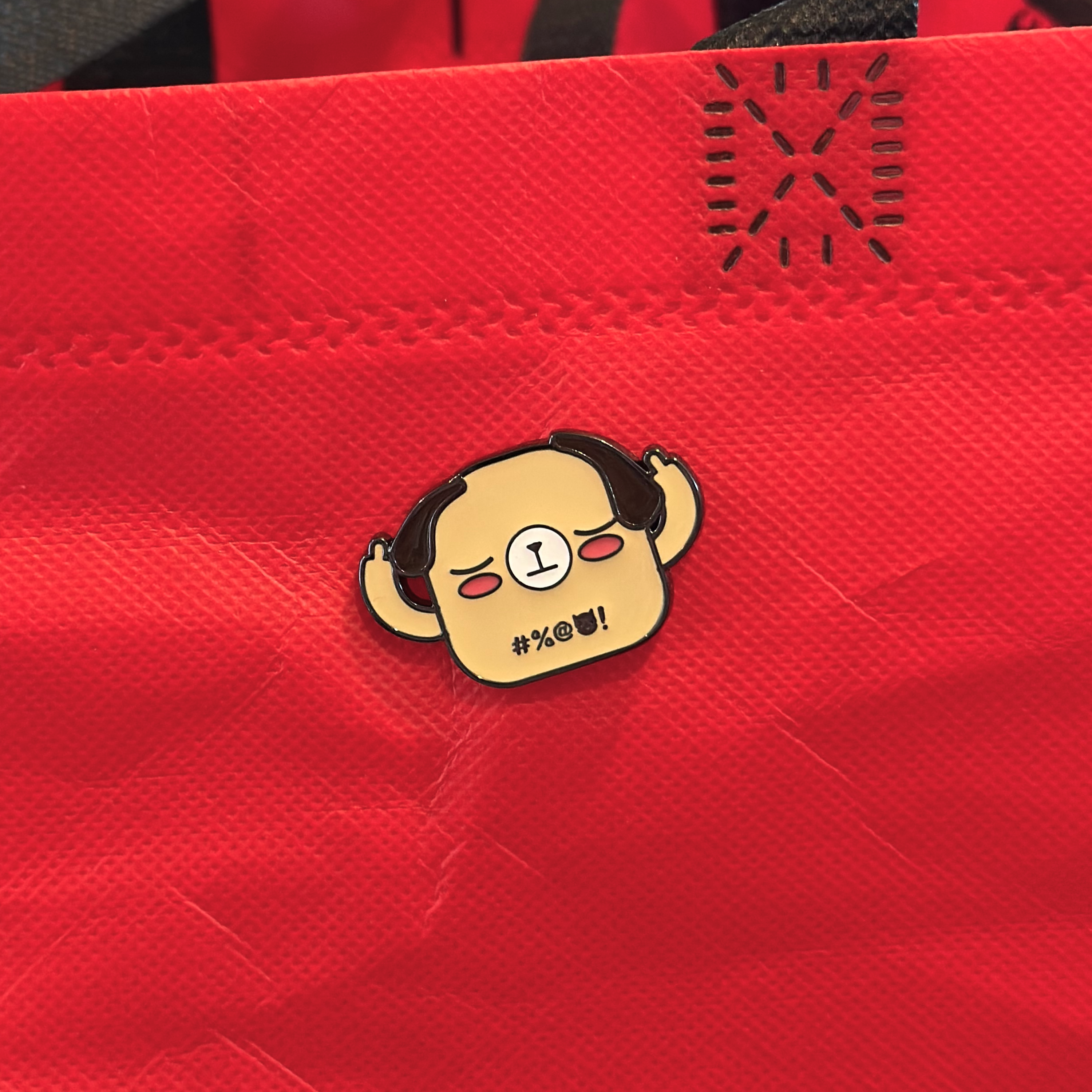 Beige dog enamel pin, middle fingers up, on red tote bag