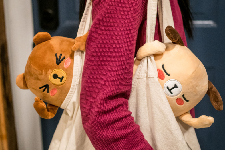 Bernard and Kai plush hanging out of a canvas tote bag carried by someone in a red sweater
